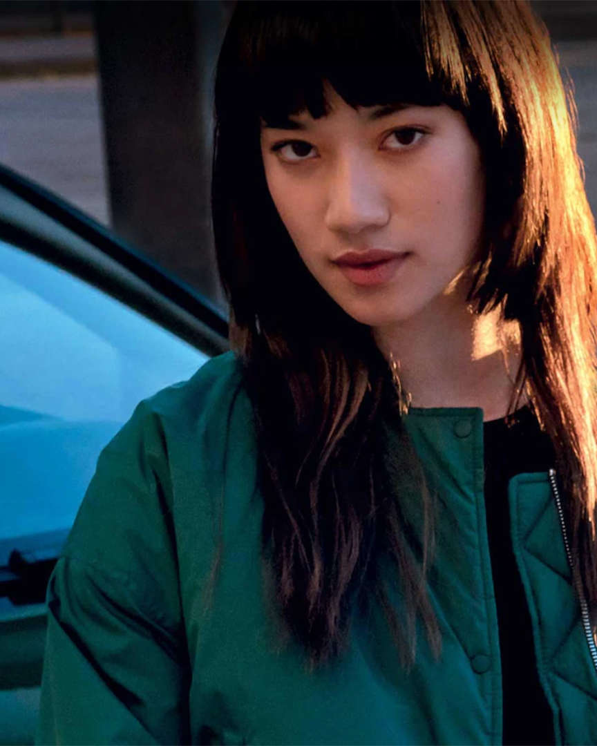 Дівчина в зеленій куртці стоїть перед автомобілем Peugeot